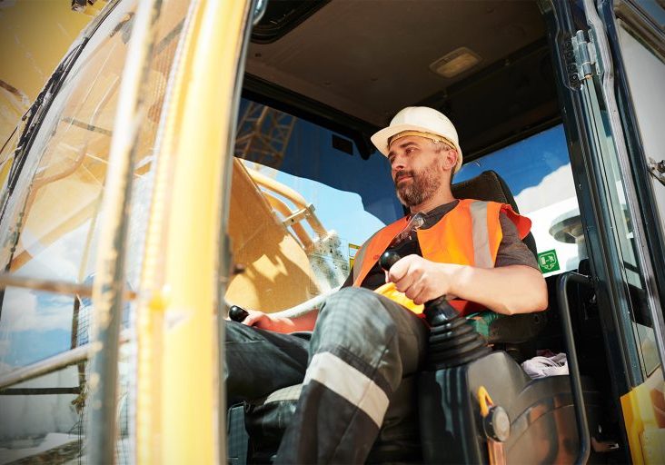 Male builder or driver of construction machine in workwear and hardhat operating caterpillar truck while sitting in cab during work